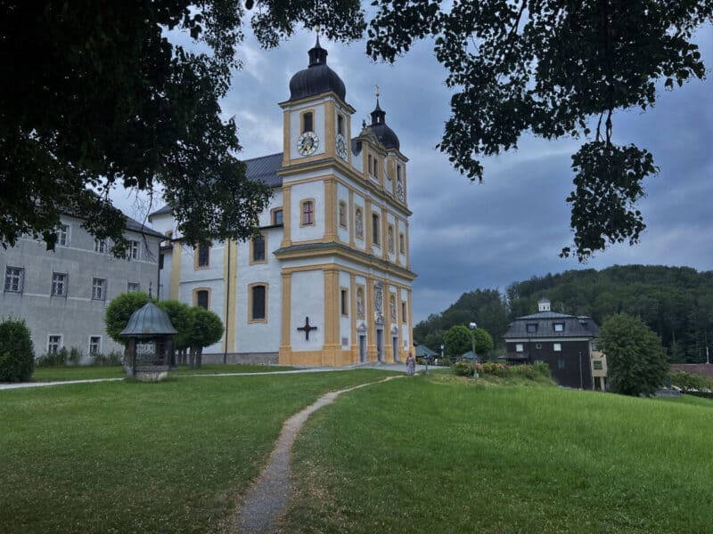 Yellow baroque-style Maria Plain church with twin towers seen from a green hilltop on the hiking trail near Salzburg
