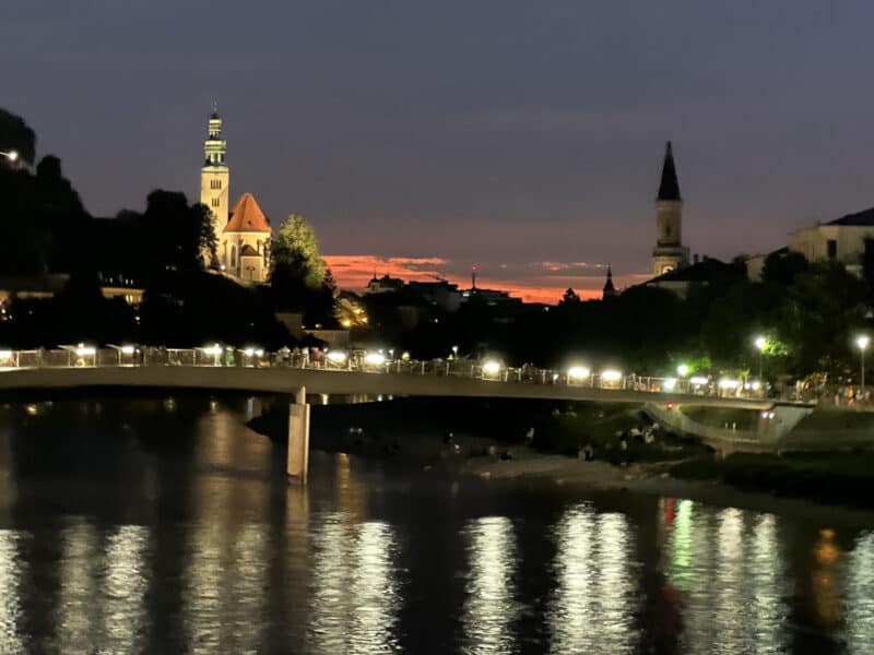 Night view of Salzburg with illuminated bridge, Salzach river reflections, and two church towers against an orange sunset sky