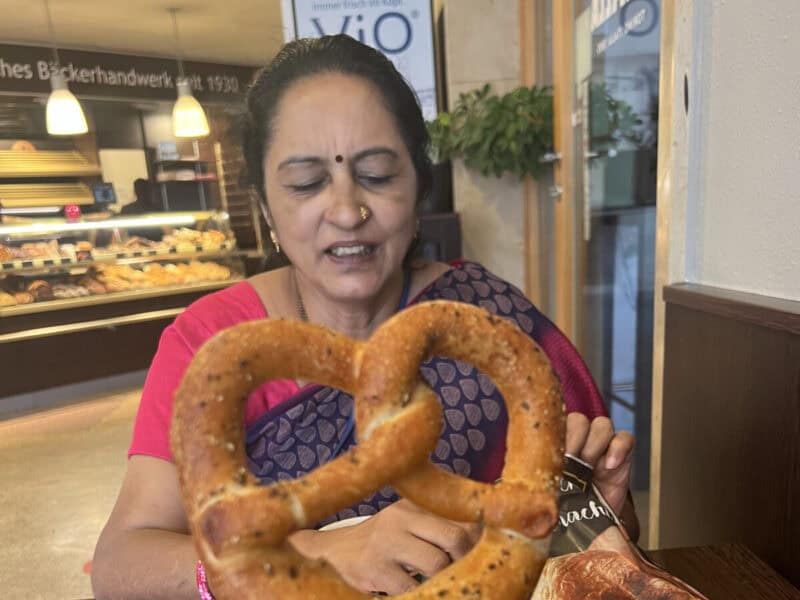 Smiling woman in a sari holding a fresh pretzel inside a traditional German bakery filled with breads and pastries.
