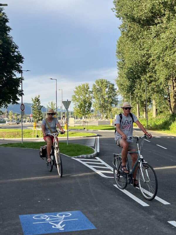 Two cyclists ride side by side on a sunny bike path in Melk, wearing hats and summer clothes, with green trees and distant hills in the background.