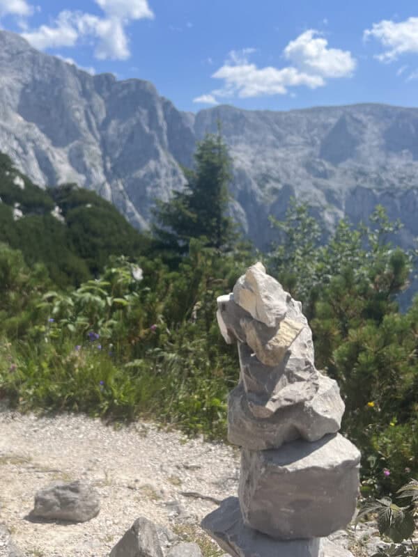 Small rock cairn stacked on a mountain trail, with blurred rocky cliffs and pine trees in the background
