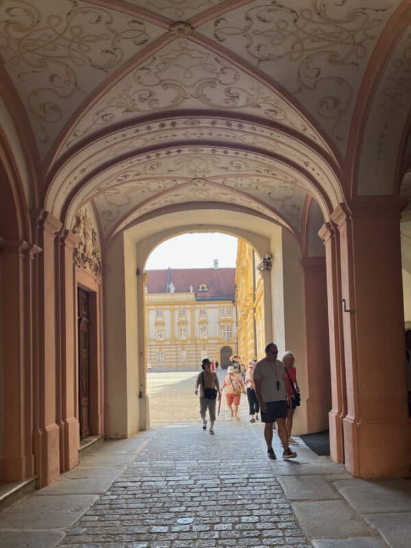 Visitors walking through an ornate arched hallway inside Melk Abbey, with pink and white ceiling patterns and bright sunlight streaming into the courtyard beyond.