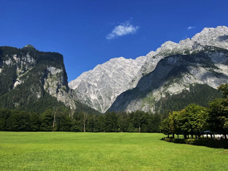 Wide green meadow at Königssee with sharp rocky cliffs and blue skies forming a striking alpine landscape.