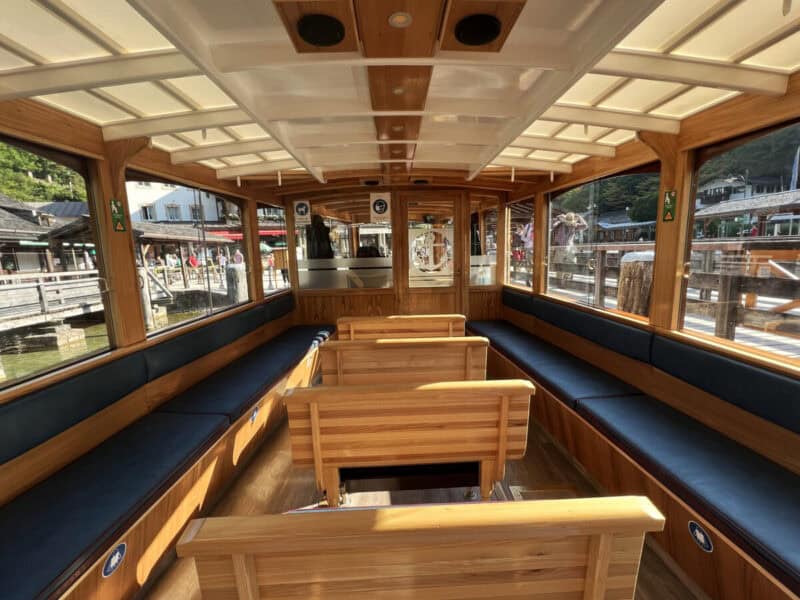 Interior of a Königssee electric boat with wooden seating, blue cushions, and reflections of lakeside shops through the glass windows.