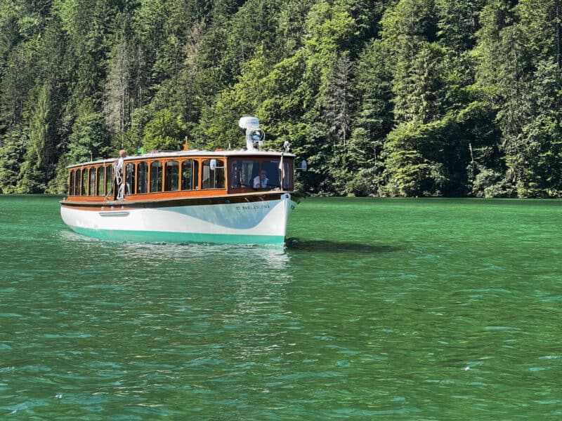 Königssee electric passenger boat cruising on emerald water with dense alpine forest in the background.