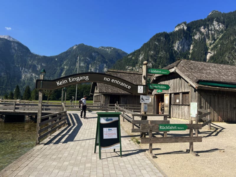 Wooden dock and signage at bathloma boat stop in Königssee, surrounded by forested peaks under a clear blue sky.