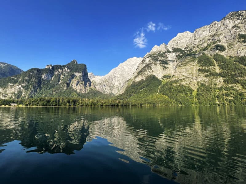 Mirror-like reflection of mountain peaks on the calm emerald waters of Königssee under a bright blue sky.