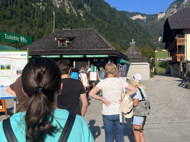 Line of tourists waiting at the Königssee ticket counters with mountains behind and map signs around.