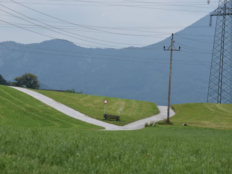 Paved trail fork on a green hill with electric poles and distant forested mountains in Salzburg countryside