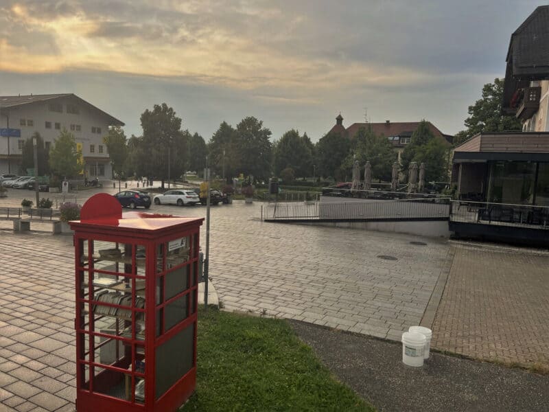 A peaceful town square at sunset featuring a classic red British-style phone booth repurposed as a tiny public library, surrounded by modern buildings, parked cars, and empty restaurant patio seating.