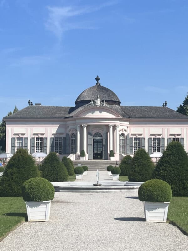Pink baroque pavilion with a domed roof, grand entrance steps, and manicured garden paths lined with clipped bushes under a blue sky.
