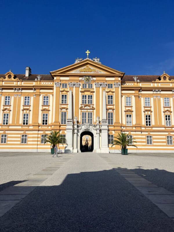 Front facade of Melk Abbey glowing in bright sunlight, featuring yellow and white baroque details, tall windows and palm trees on either side of the entrance.