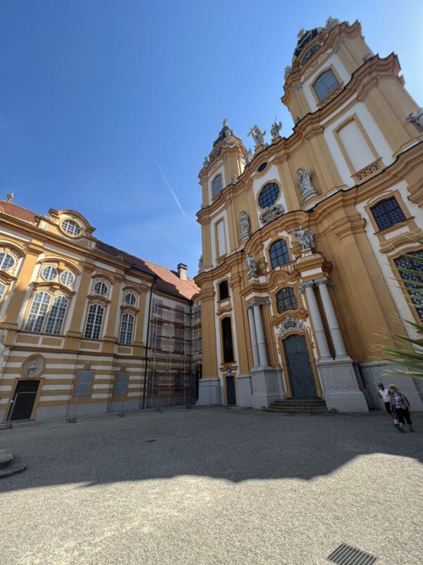 Melk Abbey’s baroque church facade in bright sunlight, with tall windows, decorative statues, and yellow and white architectural details.