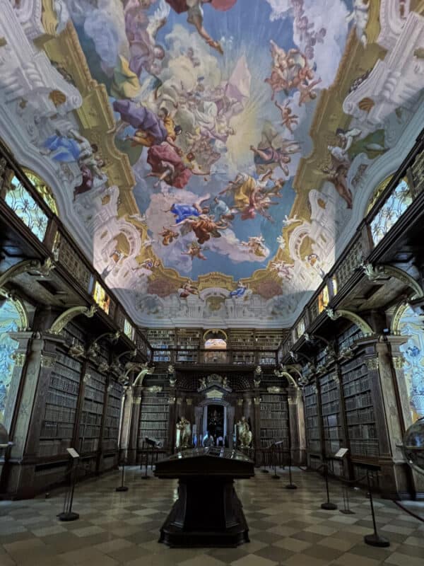 Library hall inside Melk Abbey with towering wooden bookshelves and a magnificent ceiling fresco filled with colourful angels and clouds.