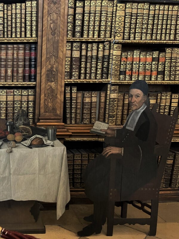 Lifelike cutout figure of a monk sitting beside a table of food, surrounded by rows of old books in Melk Abbey’s library.