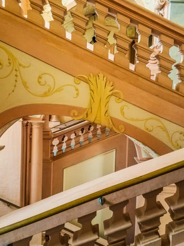 Baroque staircase inside Melk Abbey, with soft peach walls, intricate yellow scrollwork, and carved balustrades creating geometric patterns.
