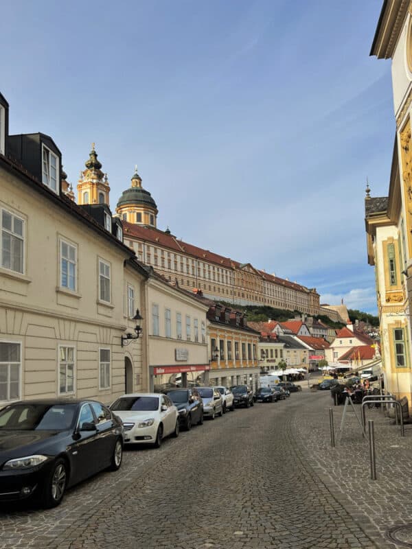 Street in Melk town lined with parked cars and pastel buildings, leading uphill towards Melk Abbey’s long baroque structure and domes under blue sky.