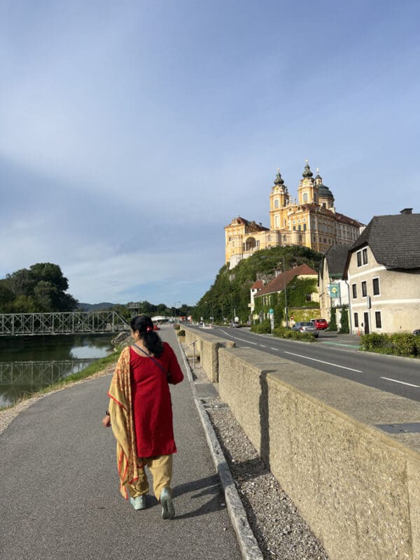 Woman in a red and beige salwar kameez outfit walking on a riverside path towards Melk Abbey, towering on a rocky hill under a blue sky.