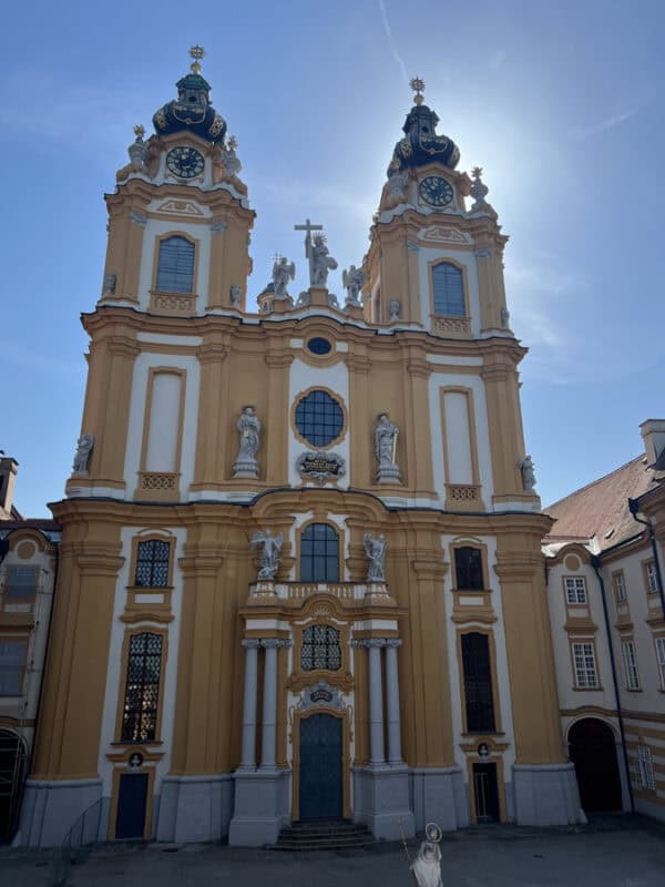 Full view of Melk Abbey’s church facade with two tall towers, statues, and intricate baroque details, bathed in sunlight under a clear blue sky.