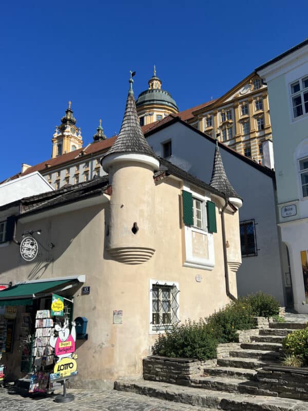 Small historic building in Melk with two round turret towers, rustic beige walls, green window shutters, and Melk Abbey’s towers visible behind it and steps leading to abbey