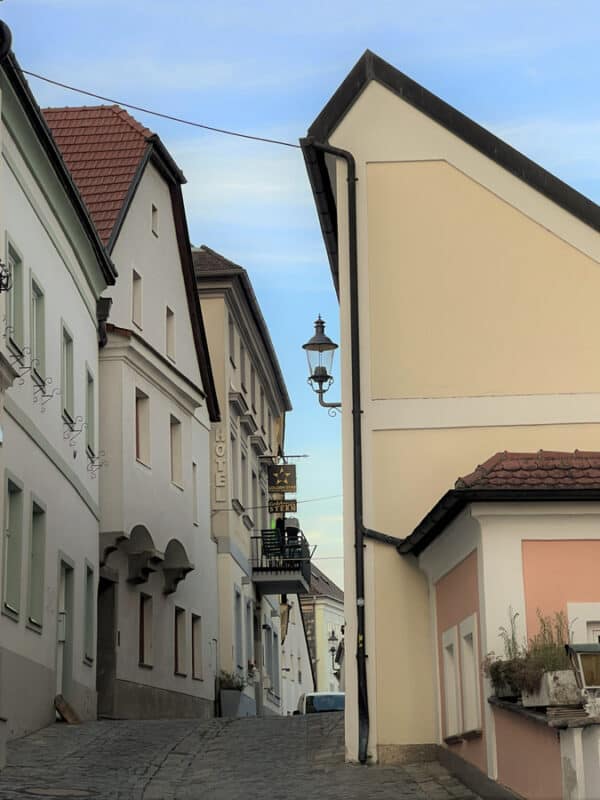 Narrow cobbled street in Melk lined with pastel buildings, hotel signs, vintage lanterns and sloping rooftops, under a pale blue evening sky.