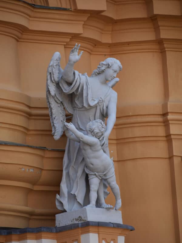 White stone statue of an angel gently guiding a child, set against the warm ochre exterior walls of Melk Abbey in Austria.