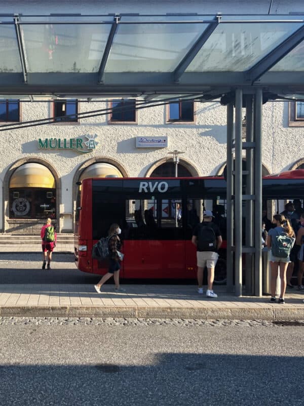 Travellers boarding a red RVO bus at Berchtesgaden station, with shops and arched windows in the background