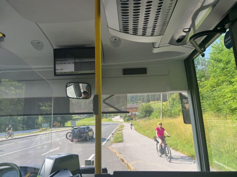Inside a public bus with a digital route display showing destinations including Fuschl am See, while outside the window, a car with bike racks drives by and a cyclist in a pink shirt rides on a parallel path.
