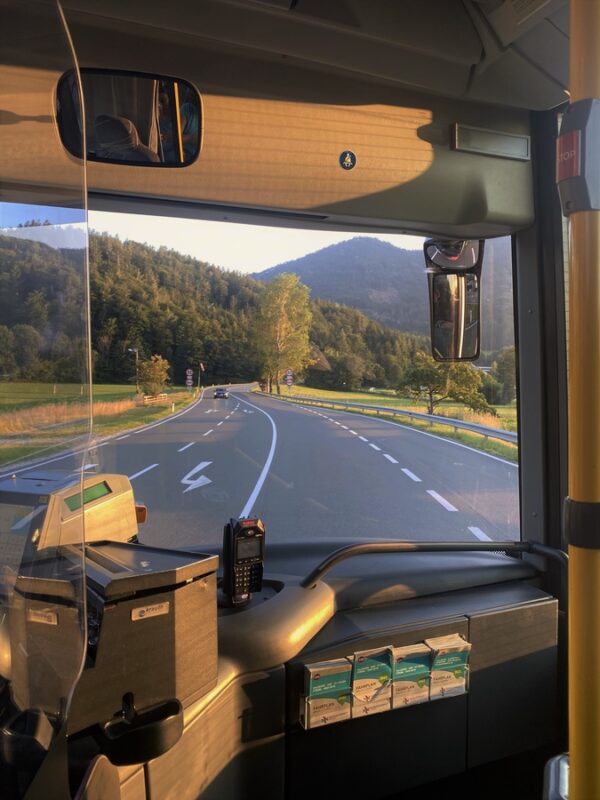 Countryside road seen from inside a public bus near Salzburg, showing card swipe machine, driver console, brochures, and a rear-view mirror