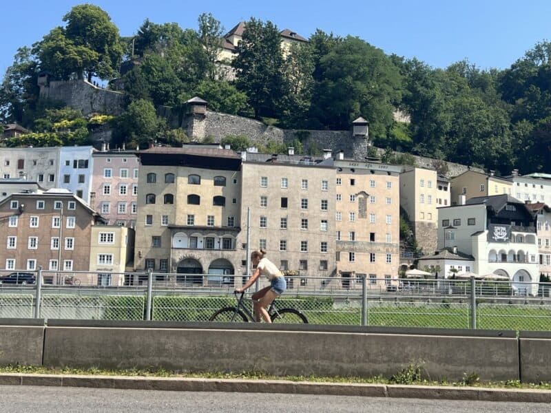 Woman cycling along the Salzach river with pastel old town buildings and city walls of Salzburg in the background