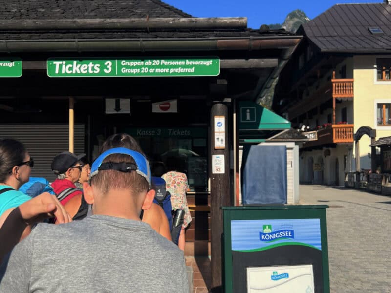 Travellers queuing at the green Königssee ticket counter in the morning light with alpine houses and blue skies in the background.