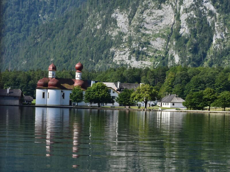 Closer view of St. Bartholomä church and surrounding buildings at Königssee, with tall green cliffs behind.