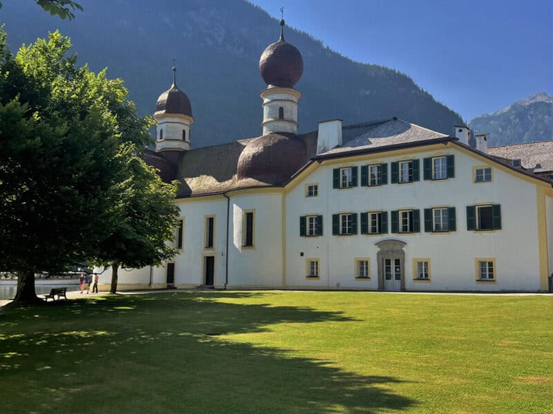 Side view of St. Bartholomä church and former royal lodge with green lawns and alpine forest backdrop.