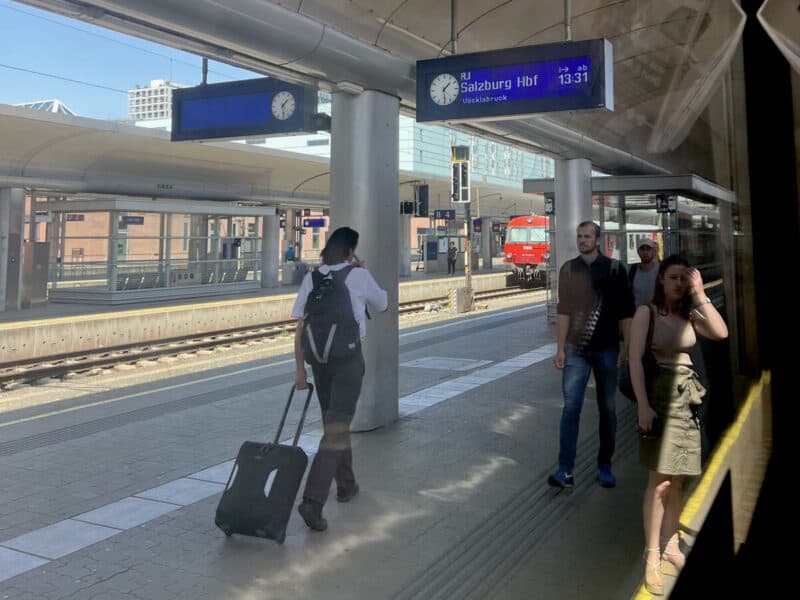 Travellers on Salzburg Hauptbahnhof platform seen from inside a train, with digital sign showing RJ train to Innsbruck