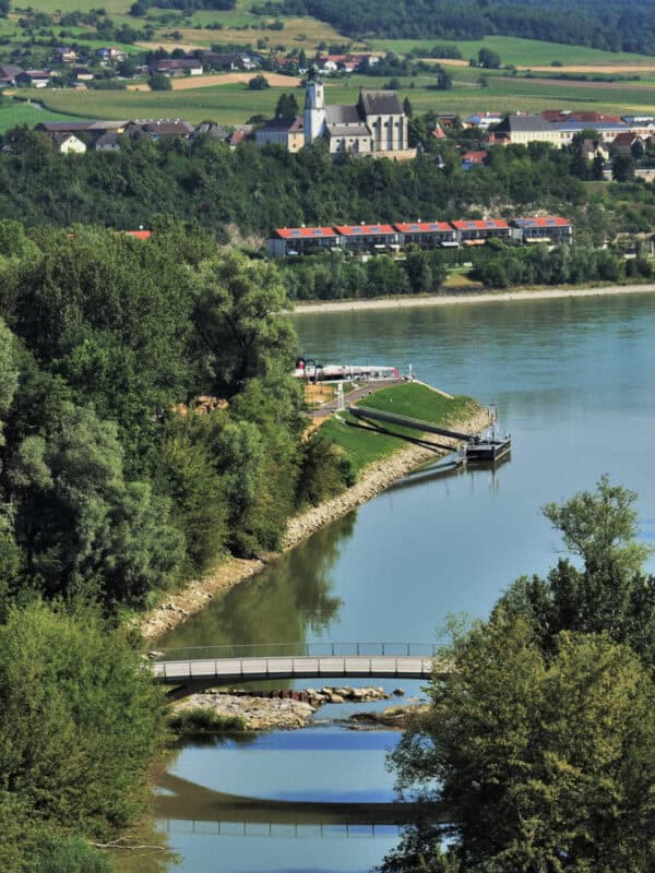 View over the Danube River with a curving bridge, green trees, and a hilltop church surrounded by red-roofed houses near Melk, Austria.