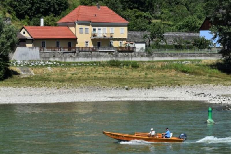 A wooden boat with two people cruising on the Danube River, with a large yellow house and stone embankment on the opposite bank in Wachau Valley, Austria.