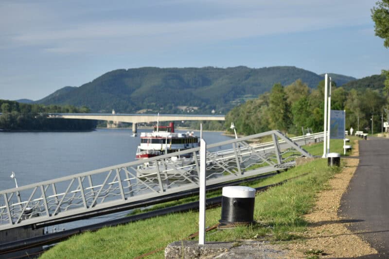 A Danube cruise ship docked along the riverbank in Wachau Valley, Austria, with a bridge and green hills in the background under soft morning light.