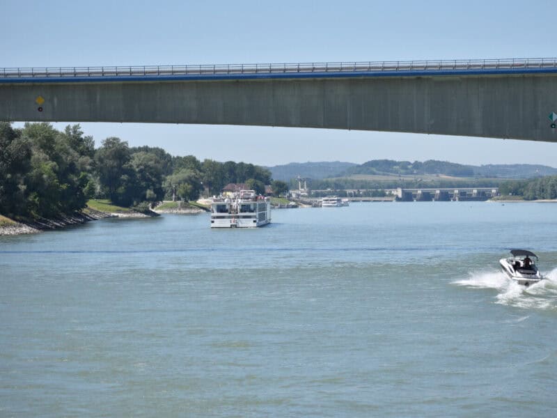 A concrete bridge spans the Danube River, with a white river cruise boat sailing underneath and a speedboat creating ripples in the foreground, distant hills visible beyond.