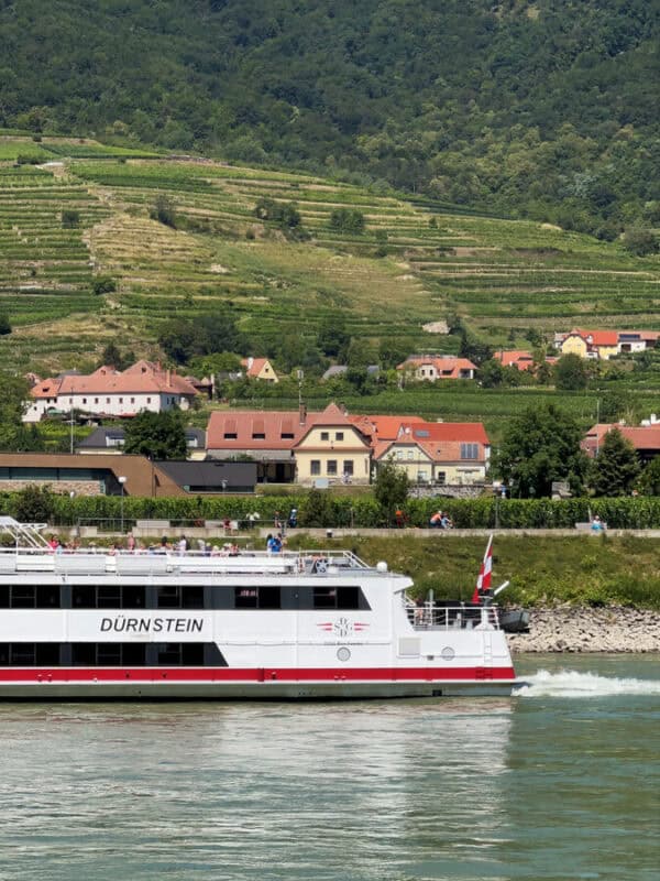 White Danube cruise boat named Dürnstein sails past vineyards, terraced green hills, and red-roofed houses in the Wachau Valley, Austria, under bright summer sunshine.