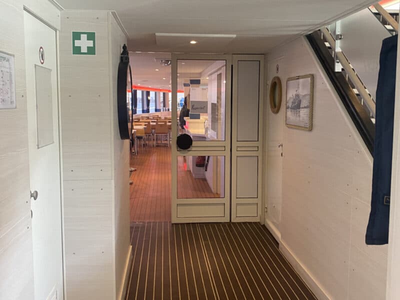 Narrow interior corridor of a Danube river cruise ship, with striped wooden floor, white panelled walls, first aid sign, stairs leading up, and a glass door opening to a dining area with wooden chairs.