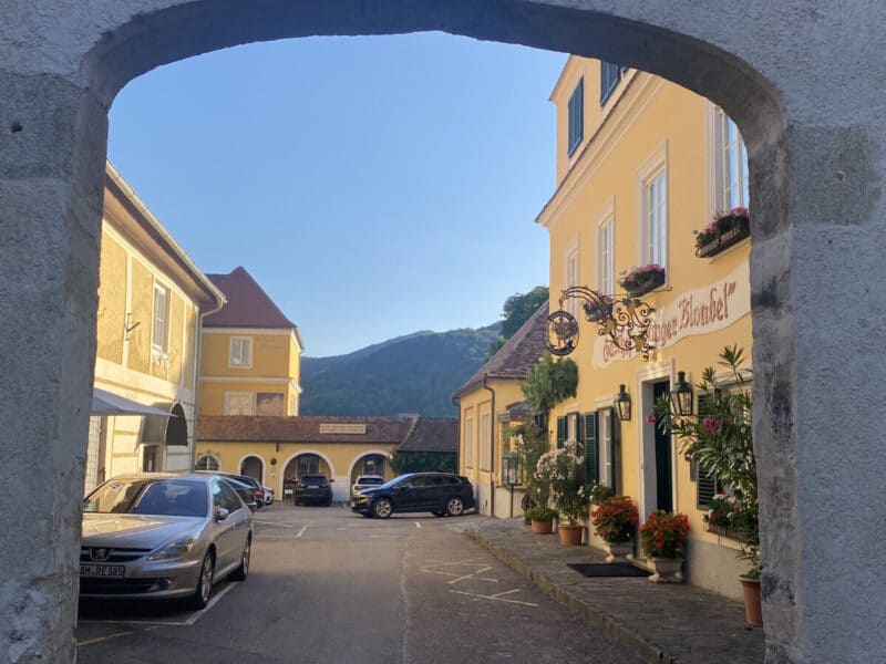 View through a stone archway towards a sunny square in Dürnstein, with yellow baroque buildings, potted flowers, and parked cars, framed by distant green hills.