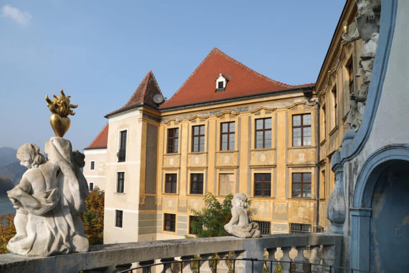 Baroque-style yellow and cream building of Dürnstein Abbey with red-tiled roofs, statues on a stone terrace, and views towards the Danube under clear skies.
