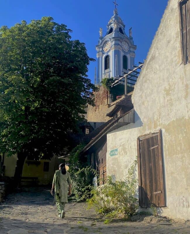Woman wearing green clothes walking on a stone path under trees in Dürnstein, with the blue tower of the abbey rising behind traditional buildings in bright sunlight.