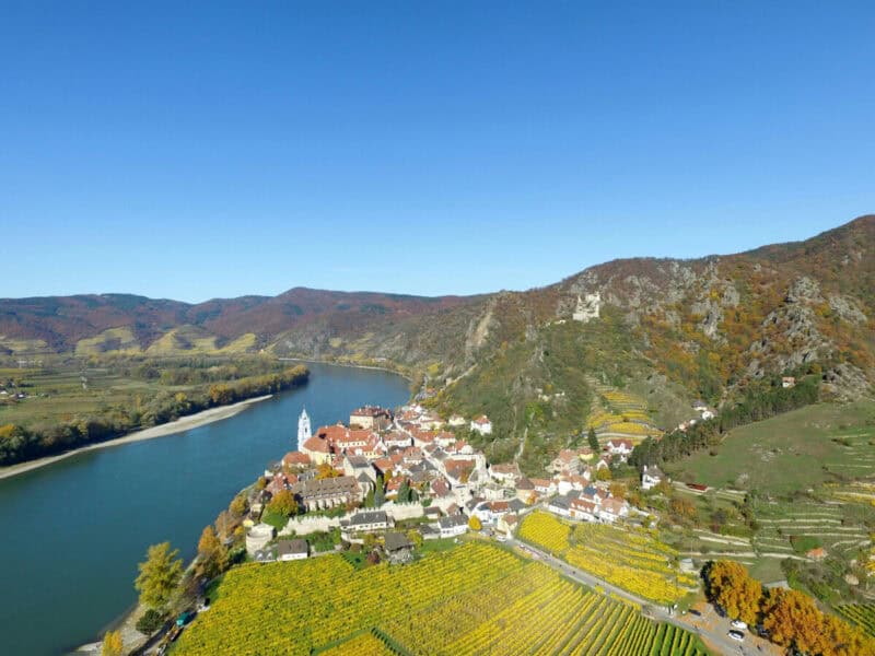 Aerial view of Dürnstein in the Wachau Valley, showing the blue abbey tower, red rooftops, yellow vineyards, and the winding Danube river surrounded by green and autumn-coloured hills.