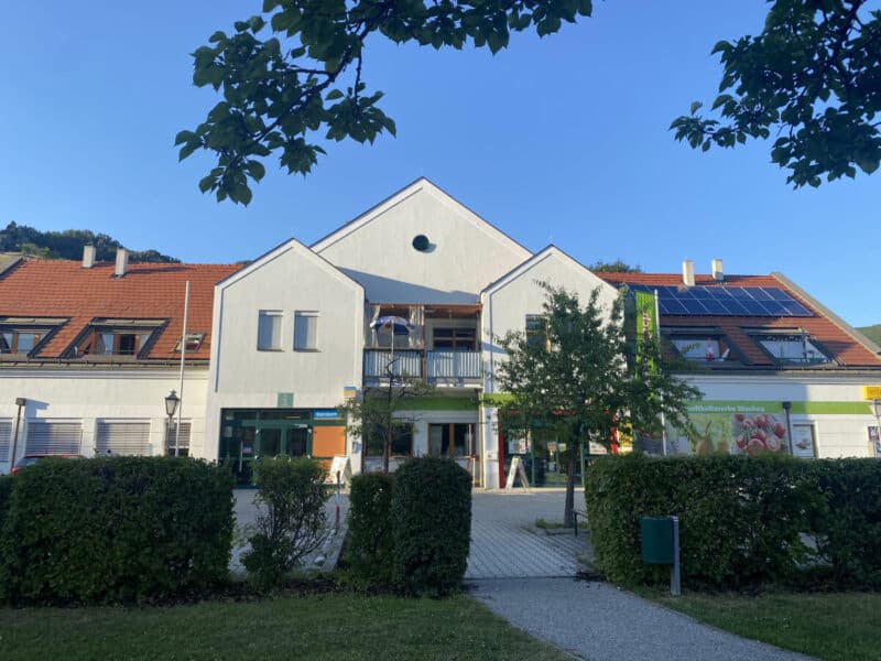 Modern white building of Nah&Frisch with red-tiled roof and solar panels in Dürnstein, framed by trees and greenery under a clear blue sky