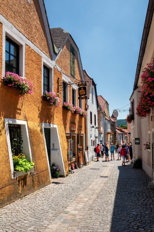 Narrow cobblestone street in Dürnstein lined with bright orange and white houses, flower boxes, and groups of tourists walking under a sunny blue sky.