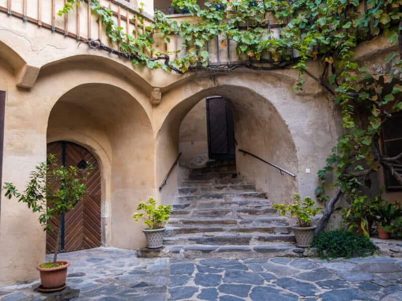 Stone courtyard in Dürnstein townhall with curved arches, vine-covered balcony railings, and a stone staircase leading to an upper wooden door, surrounded by potted plants.