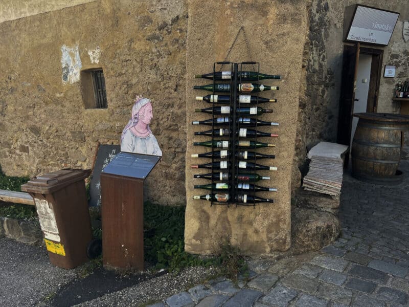 Outdoor wine bottle rack fixed to a stone wall in Dürnstein, next to a cut-out figure of a medieval woman and an entrance to a small vinothek shop.