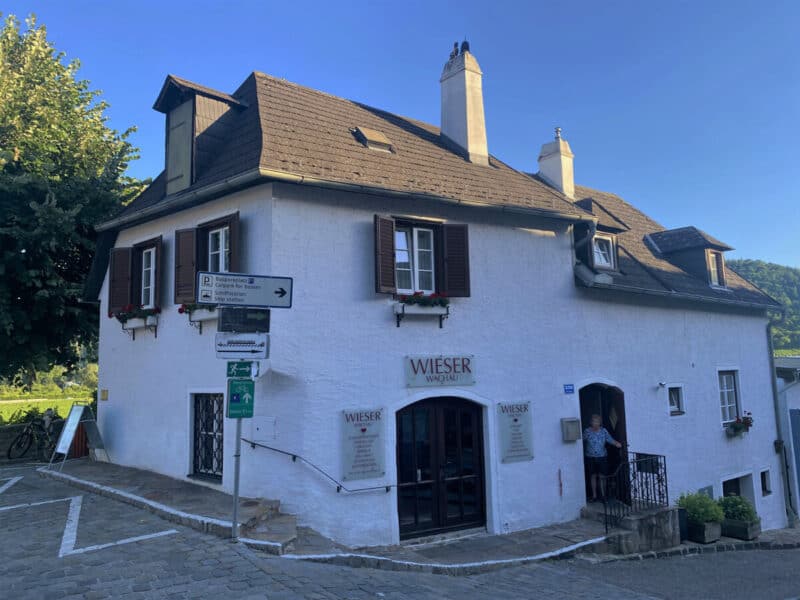 White building with dark brown roof and wooden shutters in Dürnstein, housing Wieser Wachau distillery, with signs pointing towards parking and the Danube ship station.