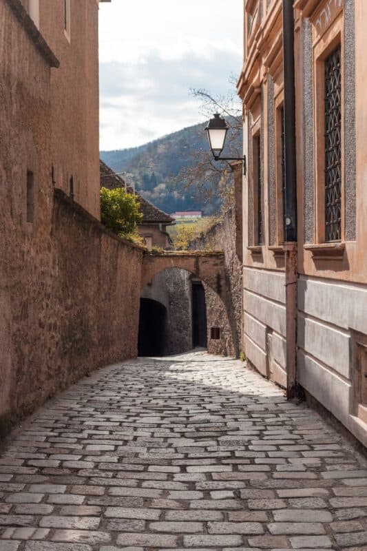 Curving cobblestone street in Dürnstein, lined with plastered buildings and an old-fashioned street lamp, with a stone archway visible in the distance and wooded hills behind.